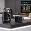 A modern kitchen countertop with a glass and stainless steel electric kettle, two black mugs, and some fruit in the background. Produktbild-Vorschau