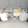 A white folding drying rack with clothes and accessories on it, set against a light gray wall and a light wooden floor. Produktbild-Vorschau