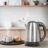 A stainless steel electric kettle is shown on a wooden countertop next to a cup of coffee and a croissant, with a kitchen background. Náhled obrázku produktu