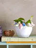 A white ceramic pot with colorful flower-like embellishments and a plant sits on top of two books. Produktbild-Vorschau