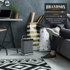 A person's legs are visible sitting on a couch next to a Brandson heater and decorative bags in a modern living room setting. Produktbild-Vorschau