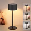 A black LED table lamp with a cylindrical shade is displayed on a desk next to a vase with flowers. The lamp is lit, casting a warm glow. The image also includes three smaller inset images showing the lamp in different settings. Náhľad obrázku produktu