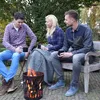 Three people sitting around a fire pit, holding mugs. Produktbild-Vorschau