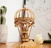 A wooden 3D puzzle of a hot air balloon sits on a wooden surface, with a blurred background of plants and a flower. Anteprima dell’immagine prodotto