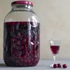 A large glass jar filled with cherries and liquid, next to a small glass of the same liquid and three cherries on a table. Produktbild-Vorschau