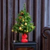 A small, decorated Christmas tree sits on a black surface in front of a dark gray curtain. Podgląd zdjęcia produktu