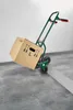 A green stair-climbing hand truck with a cardboard box on it, positioned on a concrete floor next to a step. Produktbild-Vorschau