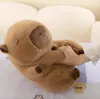 A person's hand is holding a plush capybara toy, which is lying on a surface with a floral pattern. The background appears to be a couch or soft furniture. Podgląd zdjęcia produktu