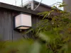 A modern outdoor wall light with a motion sensor mounted on a wooden fence, partially obscured by green foliage. Anteprima dell’immagine prodotto