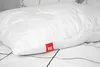 A white pillow with a quilted pattern is placed on a bed with black and white striped bedding. A red tag with a white logo and '95°' is attached to the pillow. Produktbild-Vorschau