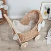 A wicker doll stroller with bedding is placed on a wooden floor, with a framed picture and a shelf in the background. Produktbild-Vorschau