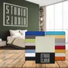 A stack of colorful fitted sheets is displayed in front of a bedroom scene, with icons and text indicating product features. Produktbild-Vorschau