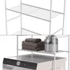 A white metal shelving unit with wire shelves, shown above a washing machine. The shelves are empty in the top portion of the image, and in the bottom portion, one shelf holds folded towels and a glass jar of detergent. Náhled obrázku produktu