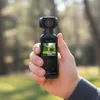 A hand holds a small black digital camera outdoors in a park setting. Produktbild-Vorschau