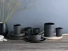 A collection of black stoneware dishes and cups arranged on a wooden table with a vase of dried branches in the background. Produktbild-Vorschau