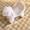 A white plush sheep rocking toy sits on a checkered rug. Produktbild-Vorschau