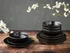 A set of black ceramic plates and bowls with gold trim, arranged on a wooden surface, with white blossoms in the background. Náhľad obrázku produktu