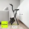 A black folding exercise bike is shown in a room next to a sofa and a white cabinet. Dimensions are indicated with text overlays. Anteprima dell’immagine prodotto