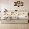 A beige upholstered bed with a built-in bookshelf headboard and a pull-out trundle bed, set in a well-lit room with decorative items on the shelves and framed pictures on the wall. Podgląd zdjęcia produktu