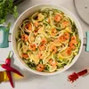 A close-up overhead view of a cooking pot filled with zucchini noodles and shrimp, garnished with lemon slices and red pepper flakes. Produktbild-Vorschau