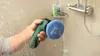 A hand is holding a green and blue Bosch Akku-Reinigungsbürste UniversalBrush, cleaning a glass shower door with soap and water. Voir les images du produit