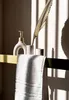 A bathroom shelf with a soap dispenser, a toothbrush holder, a dried palm leaf, and a folded towel, illuminated by dramatic sunlight casting striped shadows. Produktbild-Vorschau