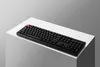 A black mechanical keyboard with a red keycap is placed on a white surface, which is on top of a cube. The background is a gradient of gray. Produktbild-Vorschau