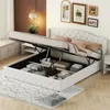 A white upholstered bed frame with the storage lid open, revealing stored items inside. The bed has a tufted headboard and is placed on a rug in a room with decorative pictures on the wall. Produktbild-Vorschau