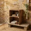 A wooden crate shelf unit is placed on a stone slab in front of a brick wall. A macrame plant hanger with ivy is on the wall to the right. A rug is on the floor. Náhled obrázku produktu