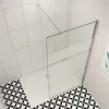 A modern bathroom shower enclosure with a glass wall and a white shower tray, set against white tiled walls and a black and white patterned floor. Produktbild-Vorschau