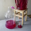 A large clear plastic carboy with pink liquid and a fermentation airlock are shown on a table. Produktbild-Vorschau