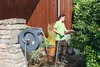 A woman is watering plants in a garden with a hose reel mounted on a stone wall. Produktbild-Vorschau