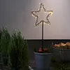 A star-shaped LED garden stake with warm white lights is placed in a pot with greenery and red apples, next to evergreen bushes and a basket of firewood. Podgląd zdjęcia produktu