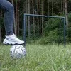 A person's leg and foot are on a soccer ball in front of a soccer goal in a wooded area. Produktbild-Vorschau