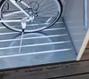 A close-up view of a bicycle's rear wheel and kickstand resting on a slatted metal floor inside a storage unit. Náhľad obrázku produktu