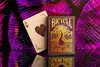 A Bicycle playing card deck and an Ace of Hearts card are displayed on a reflective surface with a vibrant, abstract purple and pink background. Produktbild-Vorschau