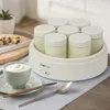 A yogurt maker with several jars filled with yogurt, next to a bowl of yogurt and fruit. Produktbild-Vorschau