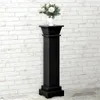 A black pedestal stand with a vase of white flowers on top, set against a white brick wall. Voir les images du produit