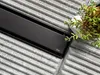 A close-up view of a black shower drain on a textured gray surface, with a small succulent plant and a white stone in the upper right corner. Produktbild-Vorschau