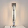 A modern LED table lamp with a metallic finish is shown on a white surface against a textured wall. Produktbild-Vorschau