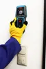 A hand wearing a yellow glove and blue sleeve holds a digital wall scanner near a light switch on a white wall. Anteprima dell’immagine prodotto