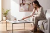 A woman is sitting on a couch in a living room, reading a magazine at a coffee table. Produktbild-Vorschau