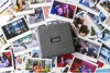A gray Fujifilm Instax LINK WIDE printer is placed on a pile of instant photos, some of which show people and outdoor scenes. Produktbild-Vorschau