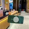 A green phone case with a magnetic charger attached is suspended in the air in front of a cafe counter. Náhled obrázku produktu