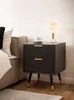 A modern black nightstand with gold accents is placed next to a bed, featuring a lamp and a cup of coffee on top. Produktbild-Vorschau