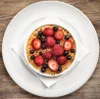 A crème brûlée dessert topped with berries, served in a white ramekin on a white plate, set on a wooden surface. Produktbild-Vorschau