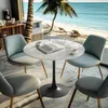 A round marble-top table with four chairs is set up in a room with a view of a beach and ocean. Produktbild-Vorschau