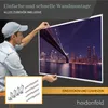 A man is holding up a framed picture of a city skyline and bridge, with mounting hardware shown in a smaller inset box. Produktbild-Vorschau