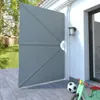 A gray folding patio side awning is partially open, attached to a white wall of a house. A backyard with a fence and trees is visible through the opening. A soccer ball and a child's scooter are on the patio. Produktbild-Vorschau