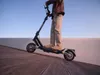 A person riding an electric scooter on a wooden deck with a clear sky in the background. Produktbild-Vorschau
